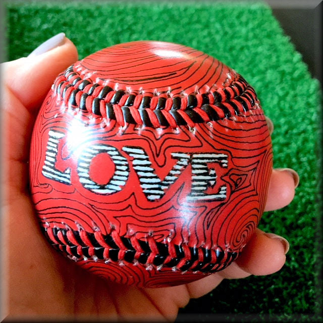 Red baseball with 'LOVE' text held in a hand against a green grass background