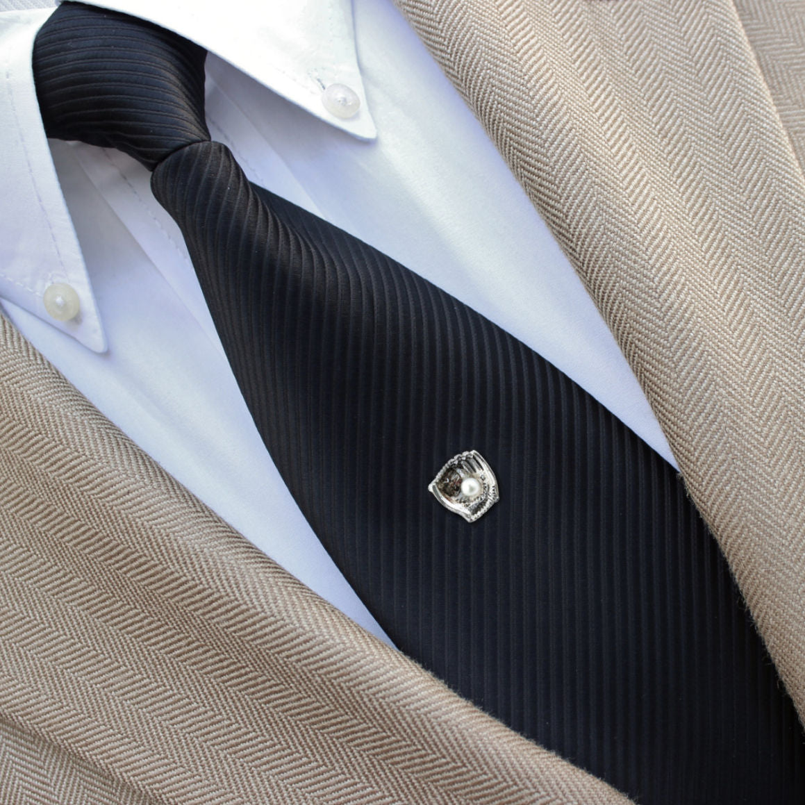 Close-up of a black tie with a decorative baseball glove tie clip, white shirt, and beige jacket.
