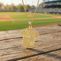Gold 'Hornets' baseball jersey necklace with number 21 on a wooden surface with a baseball field background