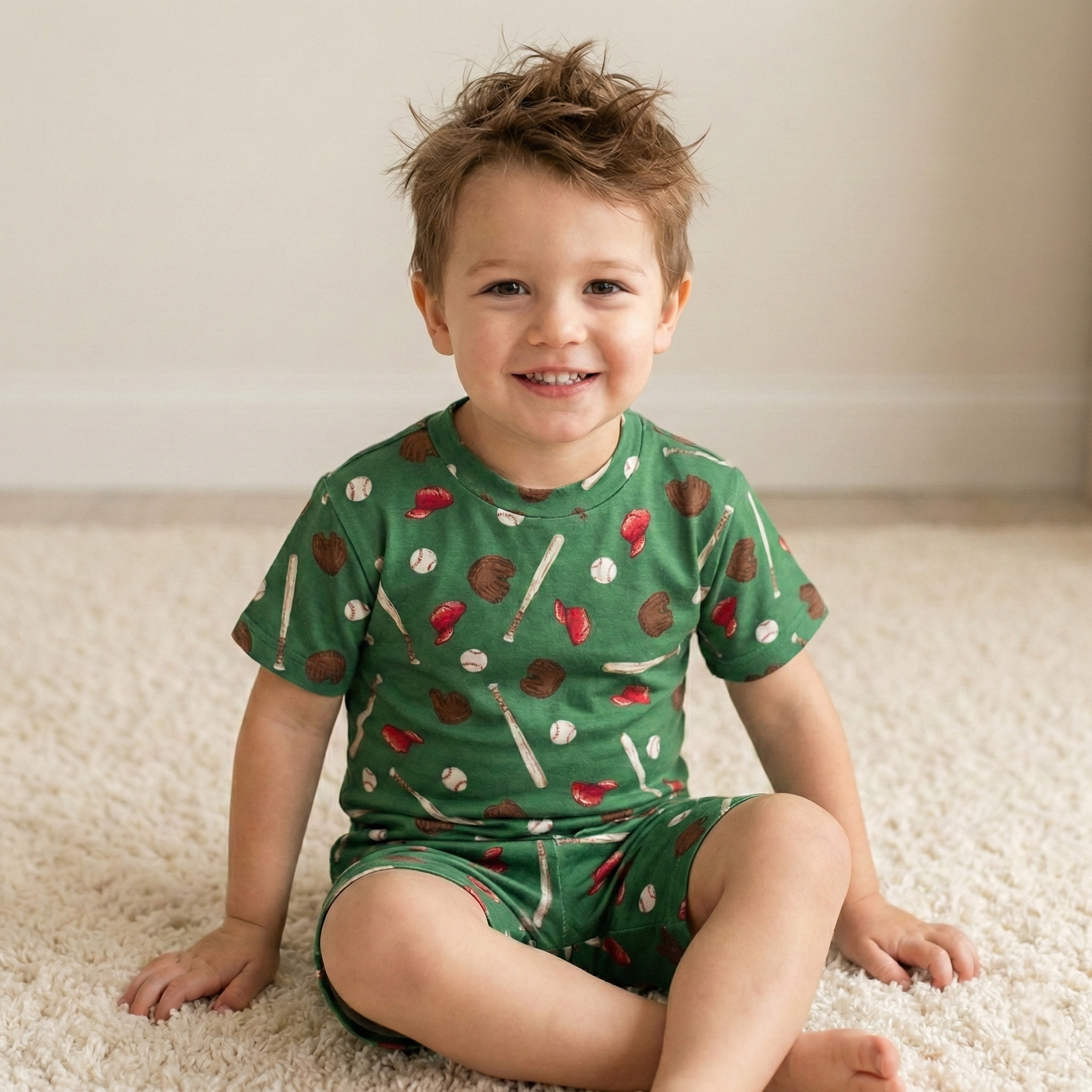 Child wearing a green baseball pajama short set with sports-themed print on a carpeted floor.