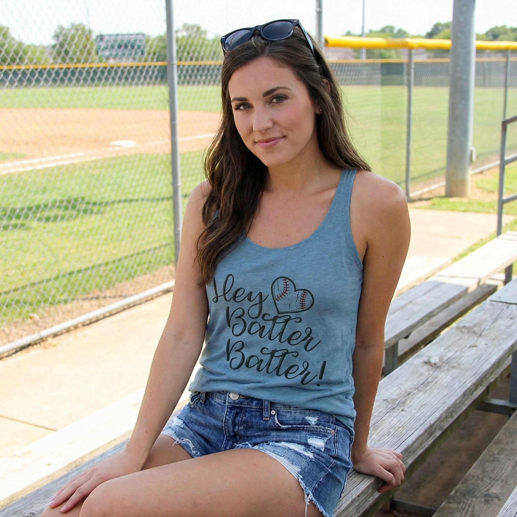 Woman sitting on a bench at a baseball field wearing a blue tank top with text.