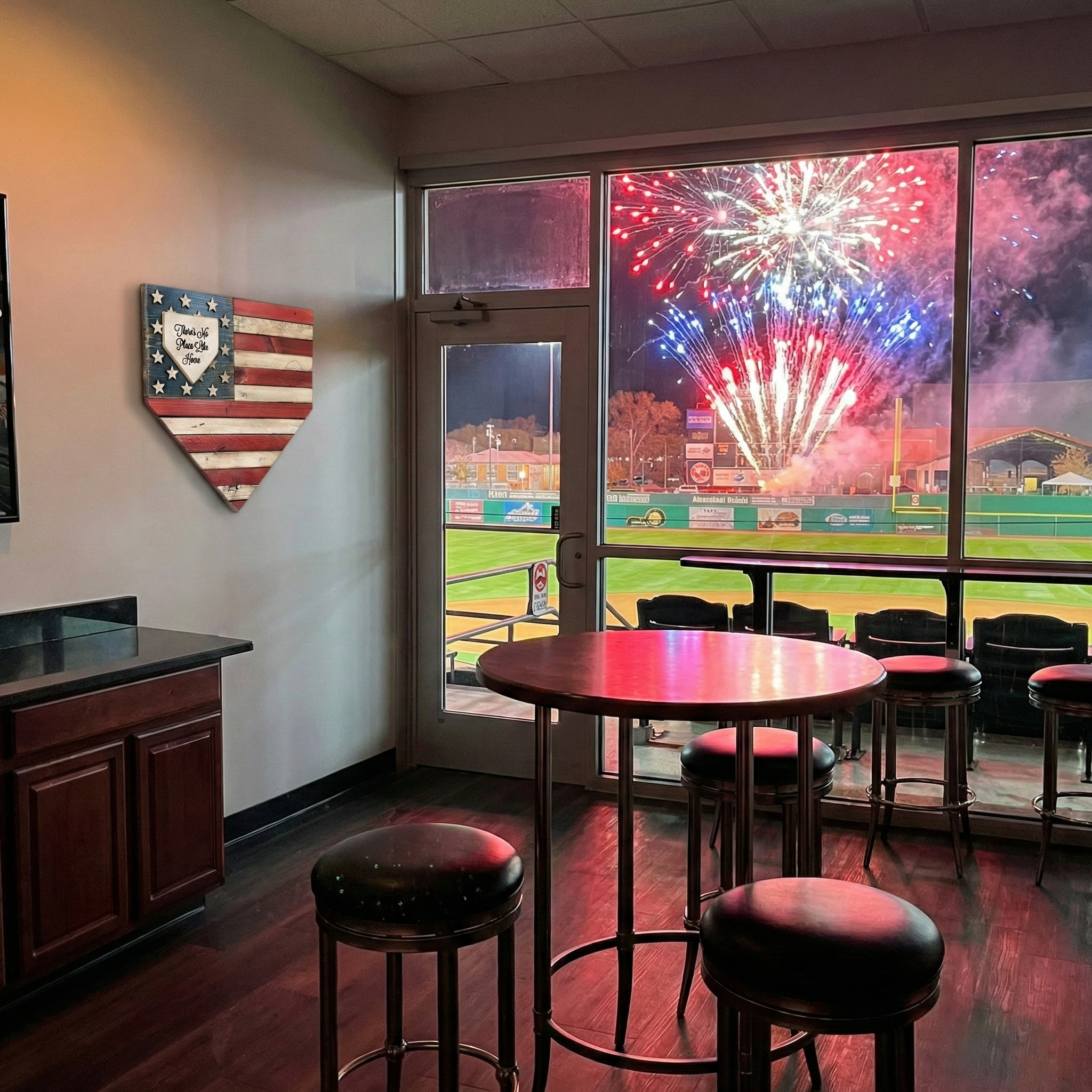 American Flag USA home plate shaped baseball wood sign in a VIP suite with a view of a baseball field and fireworks outside the window.