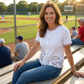 Woman sitting on a bench at a baseball field wearing a white baseball-themed shirt.