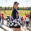 Woman holding a black tote bag with white baseball patterns at a baseball game.