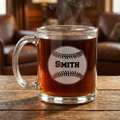 Clear glass mug with a custom engraved baseball on a wooden surface, with a blurred background of a cozy room.