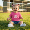 Child wearing a pink baseball shirt with 'Play Ball!' on a grassy field