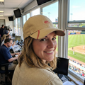 Person wearing a tan Rawlings baseball cap, sitting in a broadcast booth at a baseball stadium.