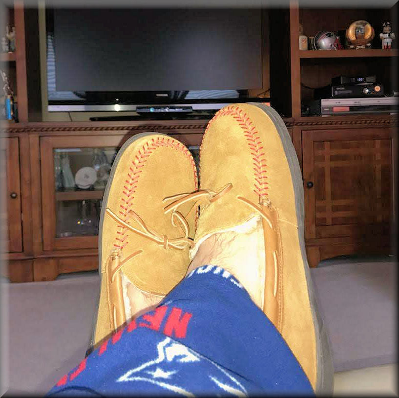 Tan suede shoes by Rawlings with red baseball stitching worn indoors, with a TV and wooden cabinet in the background.