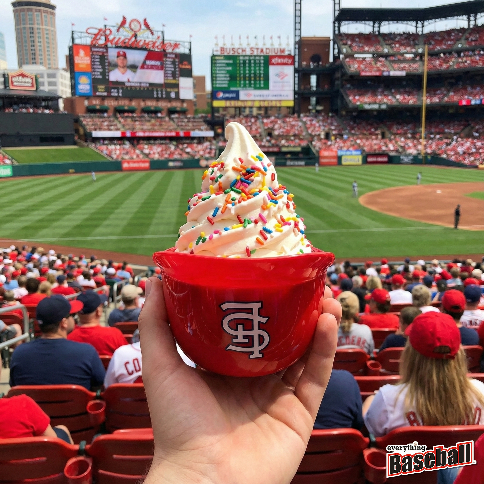 St. Louis Cardinals helmet ice cream sundae with colorful sprinkles held up at a baseball game, with stadium and fans in the background.