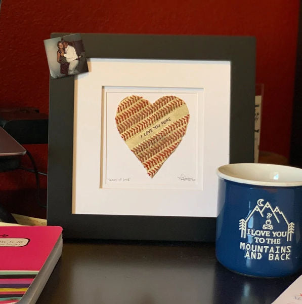 Framed baseball seams heart artwork with a mug and photo on a table