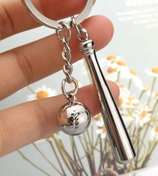 Silver keychain with a baseball bat and ball design held by a hand against a blurred natural background.