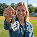 Woman holding a keychain with a baseball glove design on a sports field.