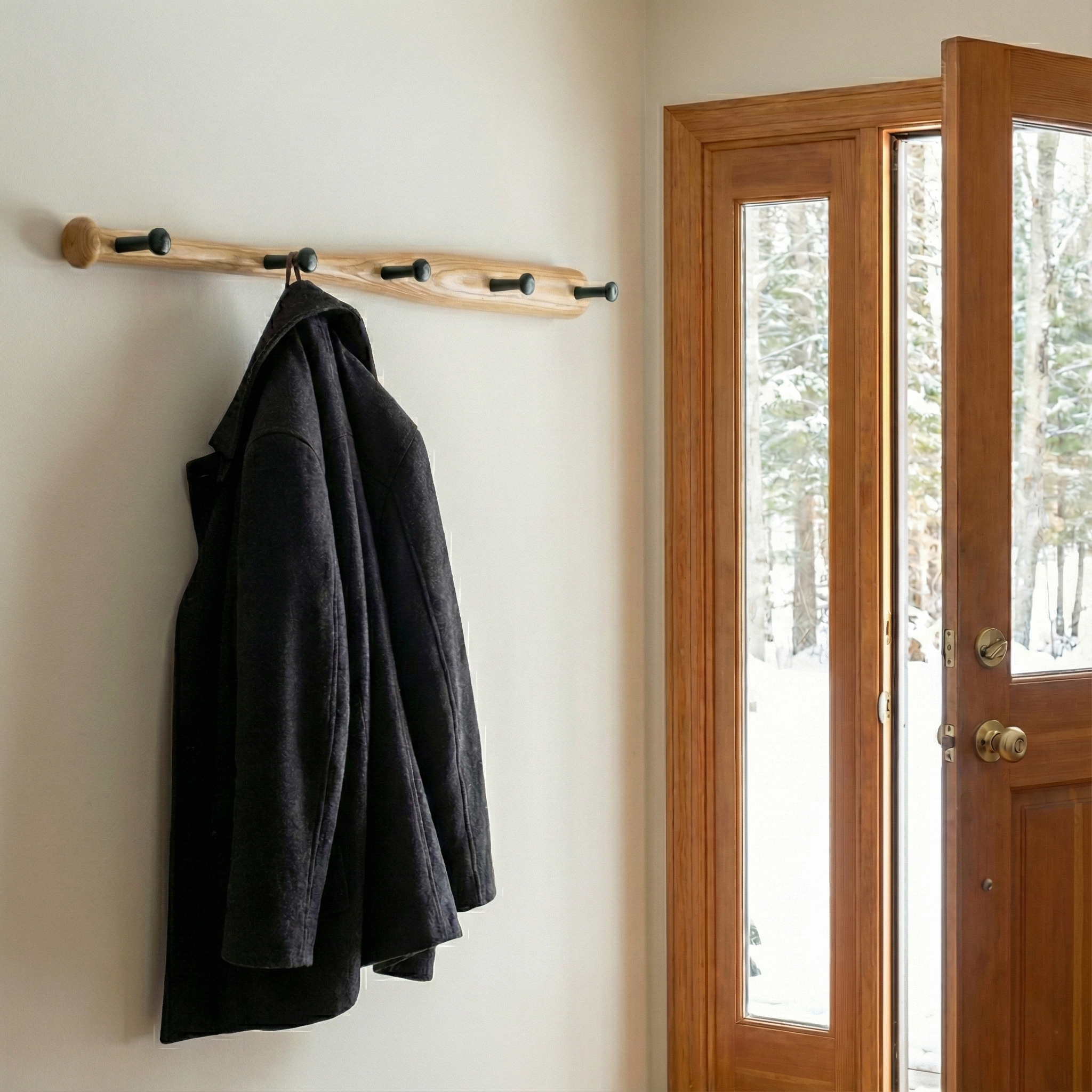 Black coat hanging on a wooden baseball bat clothes rack against a white wall with a glass door showing a snowy outdoor scene.