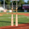Wooden baseball bat shaped earrings hanging from a hook against a blurred sports field background.